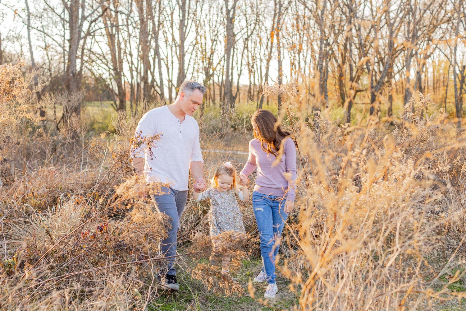 Family walking together through tall golden grass in a wooded area during sunset.