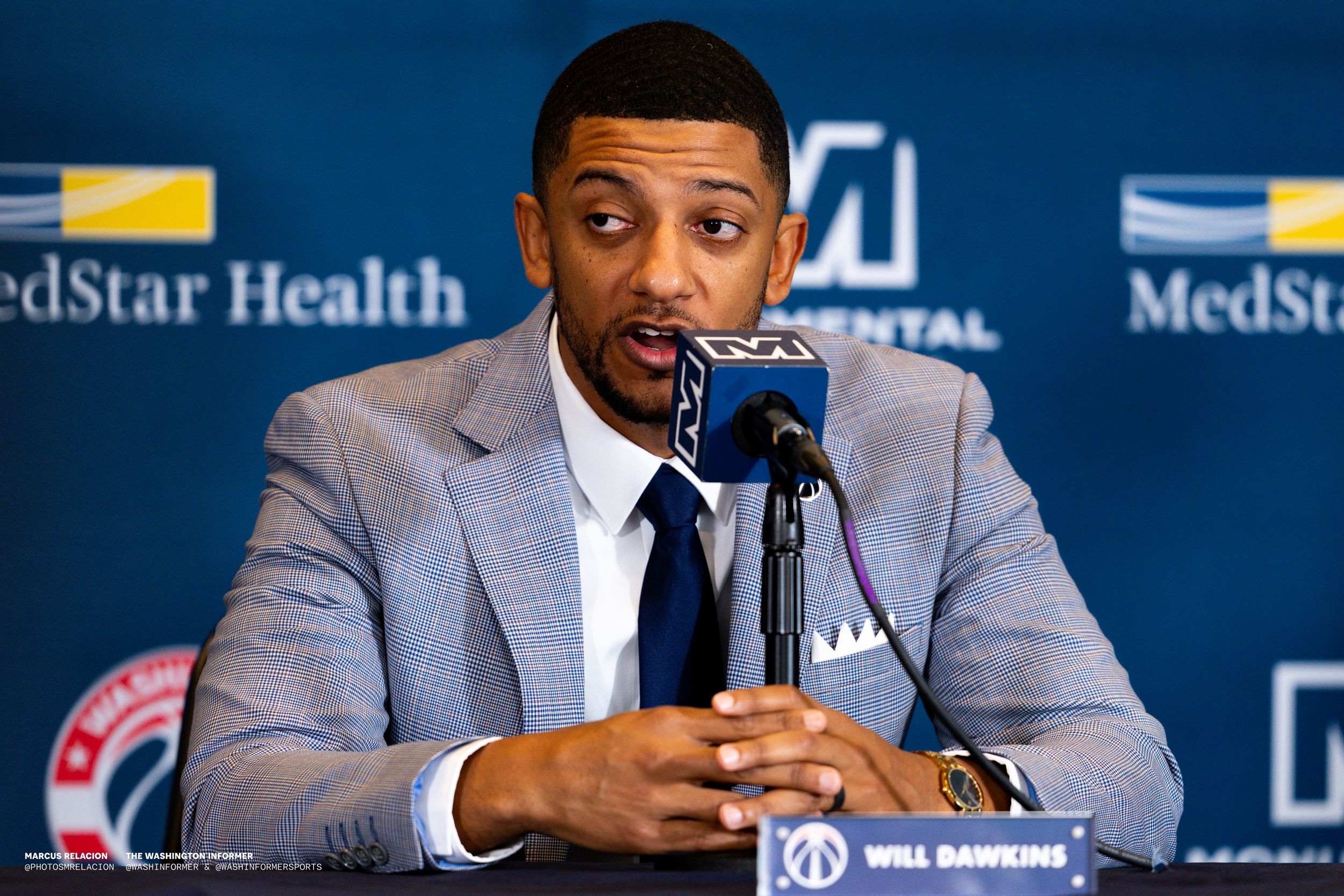 NBA basketball player holds a microphone while speaking at press conference with sponsor backdrop visible.