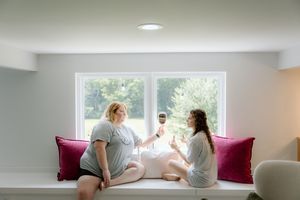 Two people sit by bright window on cushioned bench with magenta pillows, drinking red wine together.
