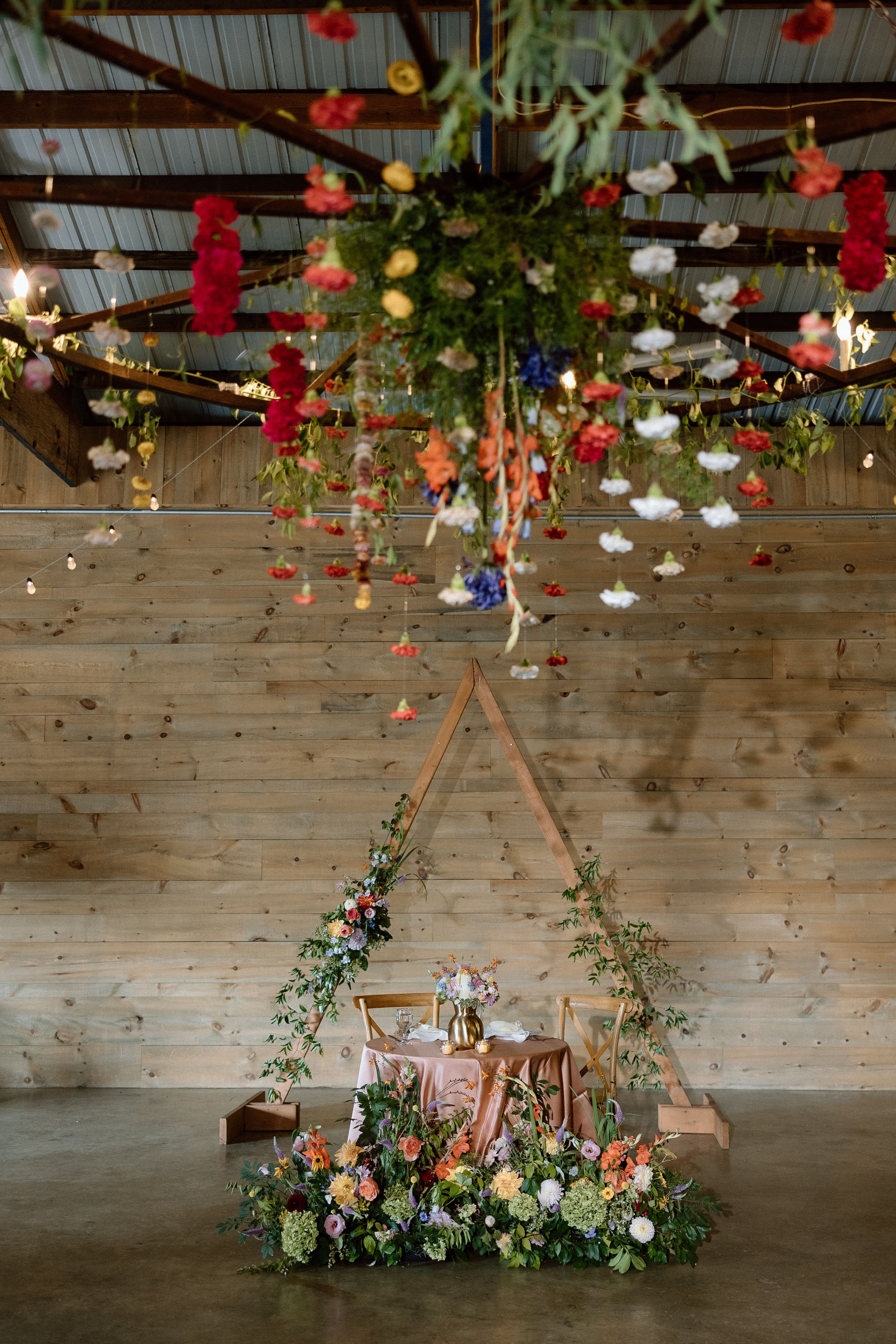 Rustic wedding sweetheart table with hanging floral installation and colorful paper lanterns against wooden wall.