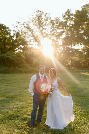 A couple stands in a sunlit meadow at golden hour with rays of sunlight streaming through trees behind them.