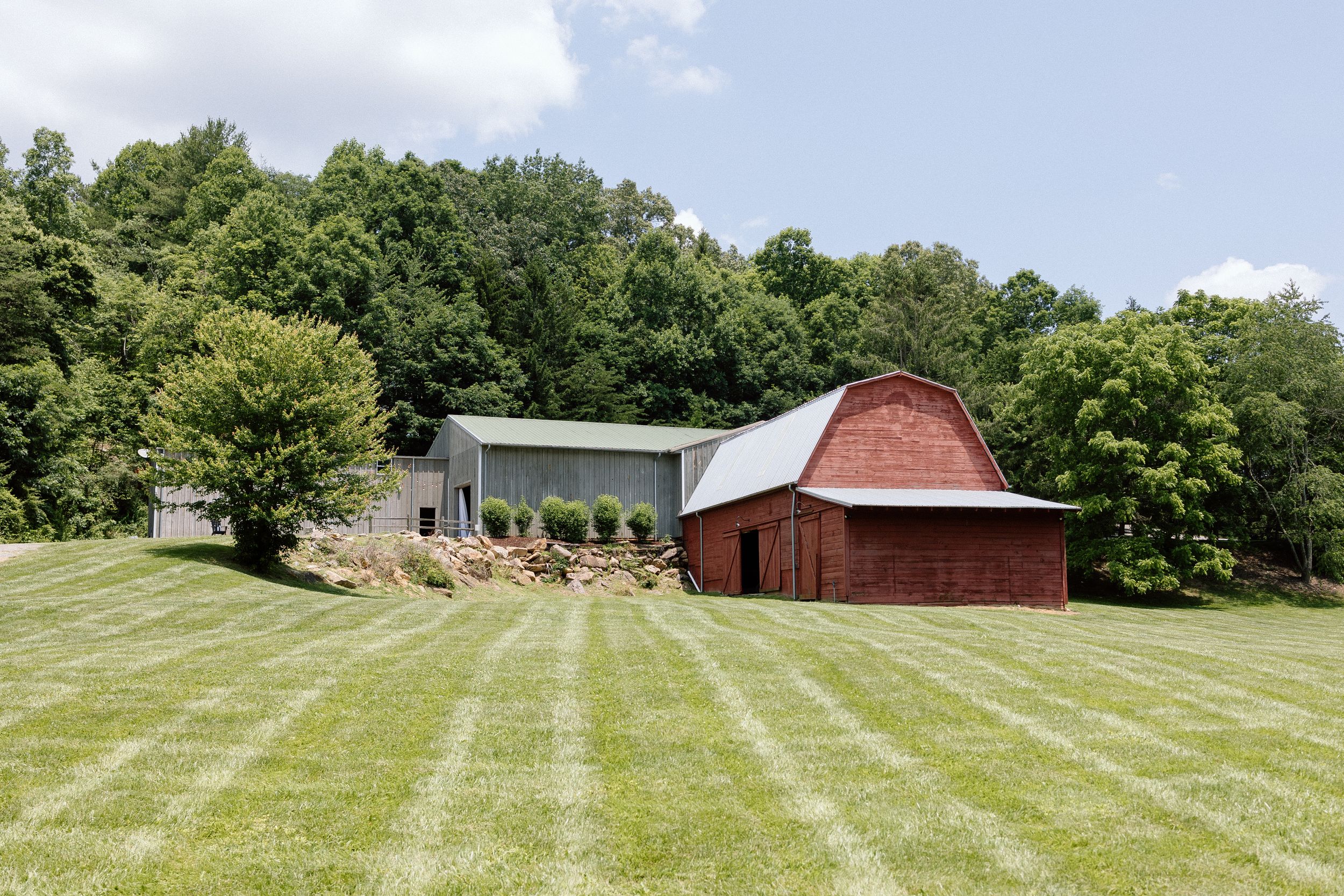 A red barn and green outbuilding sit on a freshly mowed hillside surrounded by lush summer trees.