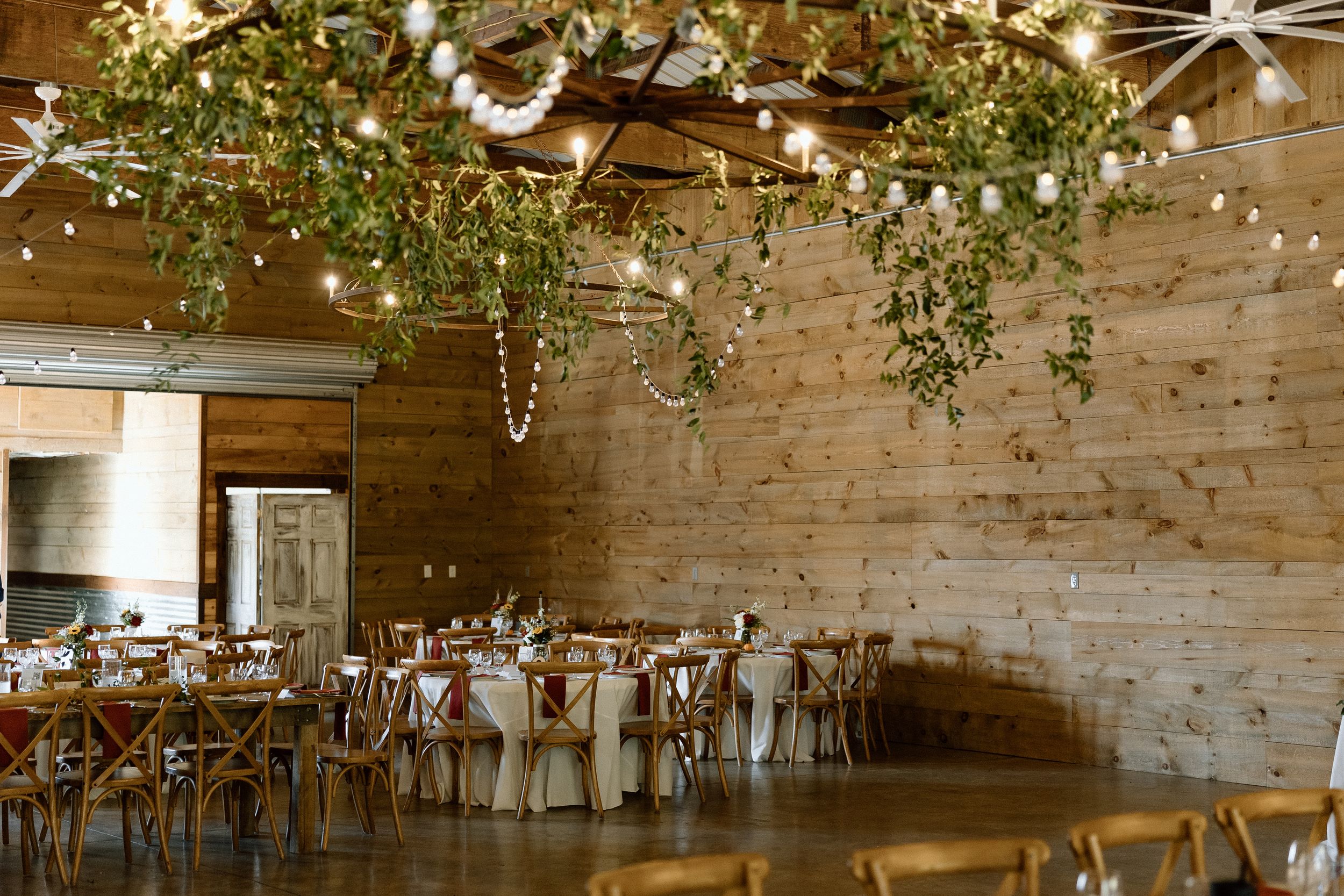 Rustic wooden barn wedding venue decorated with hanging greenery and string lights over elegant dining tables and chairs.