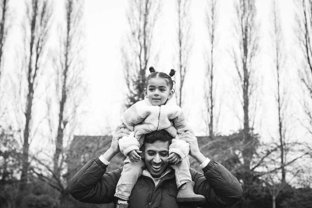 A black and white vintage photograph of a parent carrying a child on their shoulders in front of tall trees.