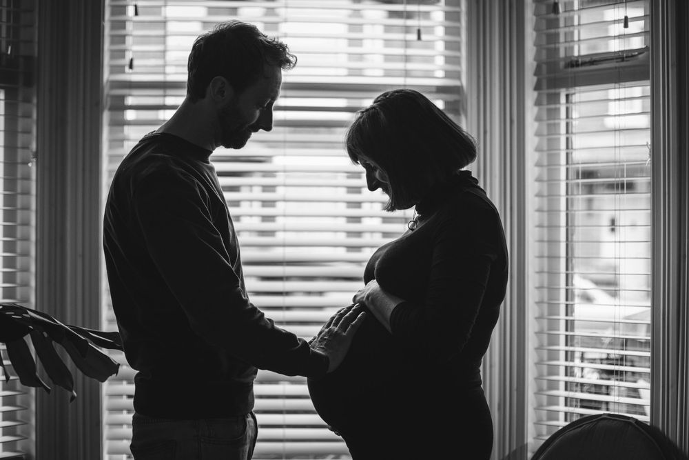 Silhouetted couple stands by window with sun streaming through blinds as they share an intimate moment.
