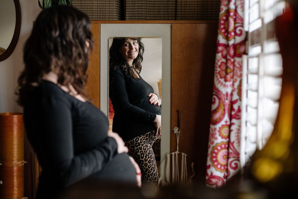 Expectant mother in dark sweater views her reflection in a mirror beside patterned curtains.