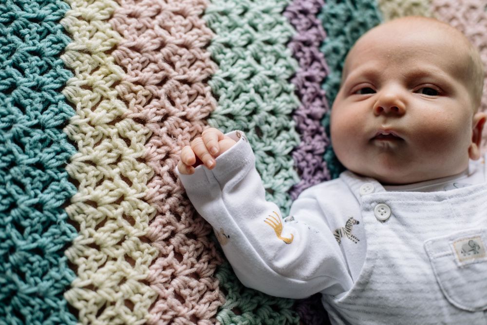 Baby lies on colorful crocheted blanket with rainbow gradient pattern in fading pastel colors.