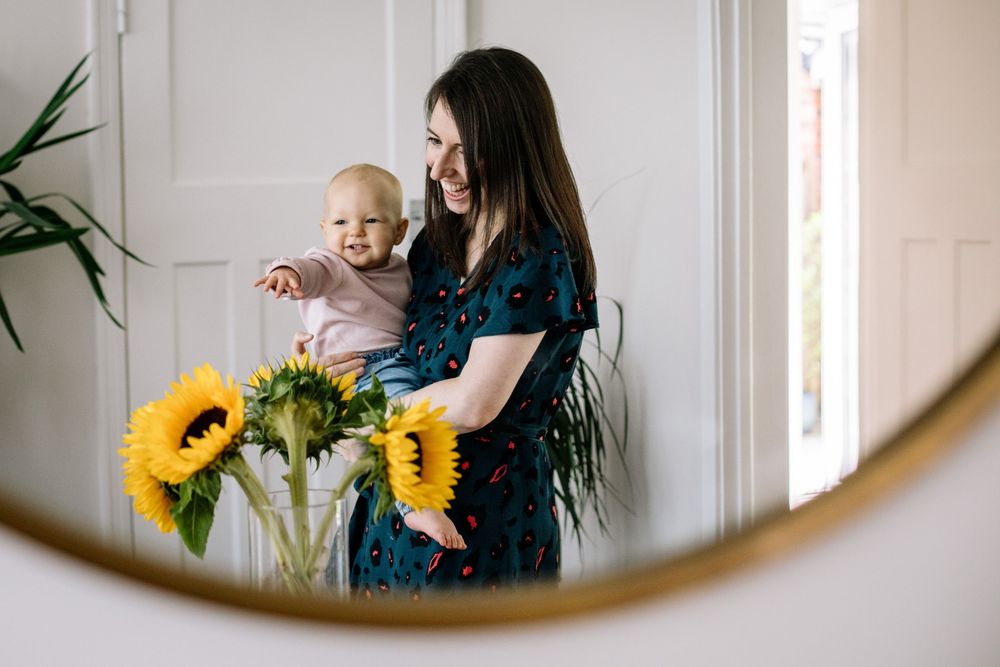 Mother in dark dress with child decorates room with sunflowers near white windows on sunny afternoon.