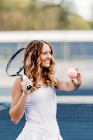 Smiling female tennis player holding a pink ball and racket on an outdoor tennis court.
