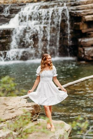 Joyful woman in white dress dancing barefoot on rocks in front of a stunning waterfall.