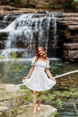 Cheerful barefoot woman in white dress posing on rocks near a beautiful cascading waterfall.