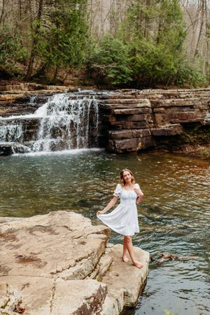 Barefoot woman in white dress twirling near a cascading waterfall surrounded by rocky terrain.