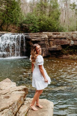 Young woman in white dress standing on rocks by a scenic waterfall in a lush forest setting.