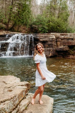 Happy young woman in white dress smiling on rocks with a picturesque waterfall backdrop.