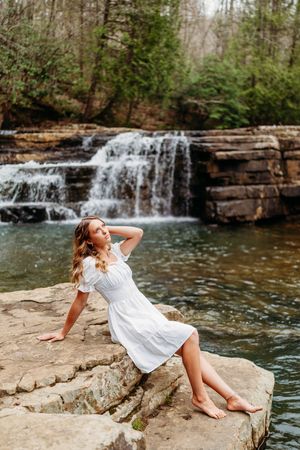 Woman in white dress reclines on flat rocks beside a wide cascading forest waterfall.