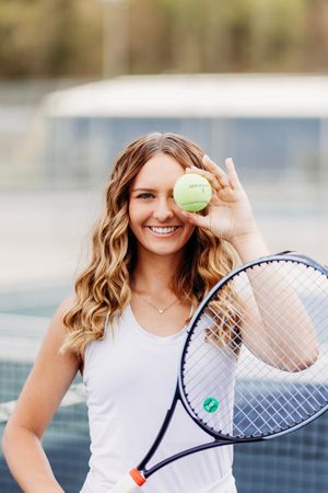 Smiling woman holding tennis ball over eye with racket on court.