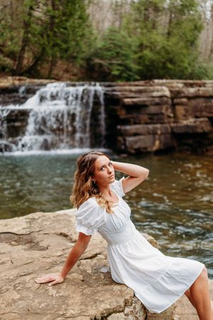 Young woman in white dress poses on rocky ledge with picturesque waterfall in background.