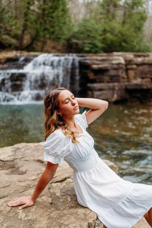 Young woman in white dress poses gracefully on rocks beside serene woodland waterfall.