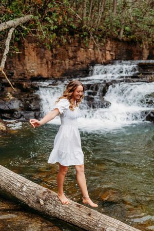 Barefoot woman in white dress walks along log with waterfall flowing over rocks behind.