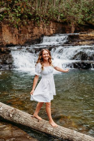Barefoot woman in white dress balances on log with scenic waterfall in background.