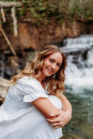 Smiling young woman in white dress poses with arms crossed near a woodland waterfall.