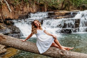 Young woman in white dress sits on log before cascading waterfall in lush forest setting.