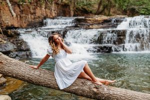 Woman in flowing white dress relaxes on log by stunning multi-tiered forest waterfall.