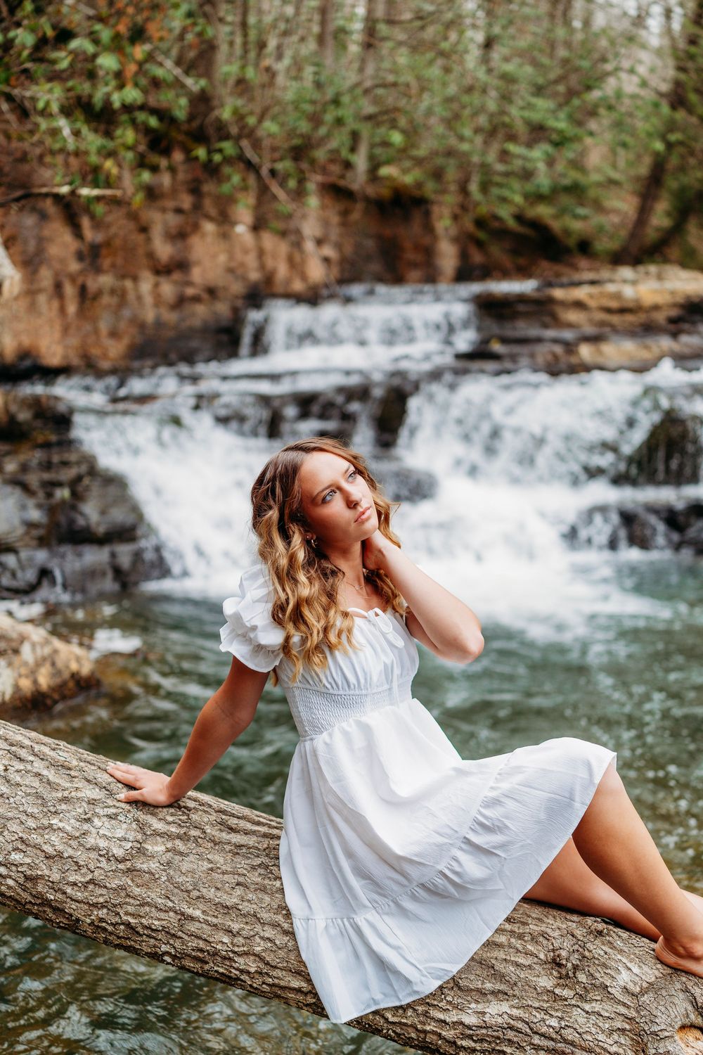 Young woman in white dress sits on a log with a serene waterfall and green forest in background.