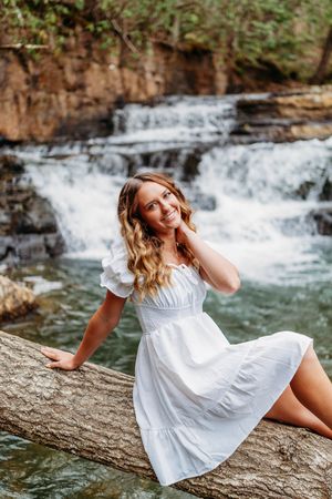 Smiling blonde woman in white dress poses on fallen log with waterfall backdrop.