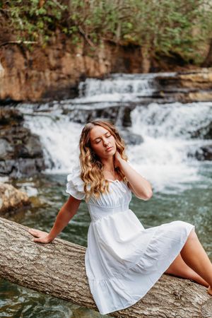 Blonde woman in white dress sits peacefully on log with cascading waterfall behind her.