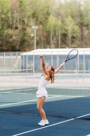 Woman serving tennis ball on outdoor blue court wearing white dress.