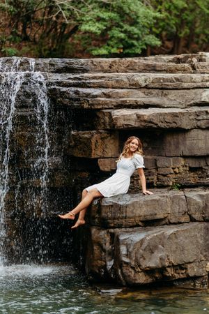 Young woman in white dress sits barefoot on rocky stone steps beside a scenic woodland waterfall.