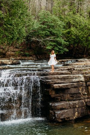 Young woman in white dress stands on rocky ledge beside a cascading waterfall in a lush forest.