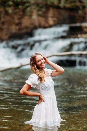Smiling young woman in white dress stands in a shallow stream with a cascading waterfall behind her.