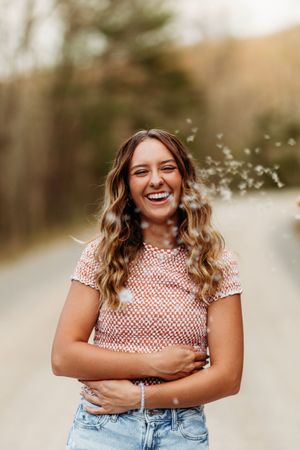 Laughing young woman in pink patterned top stands on a gravel road with confetti falling around her.