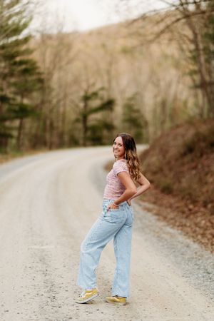 Young woman in pink top and light blue jeans poses on a winding gravel road lined with trees.