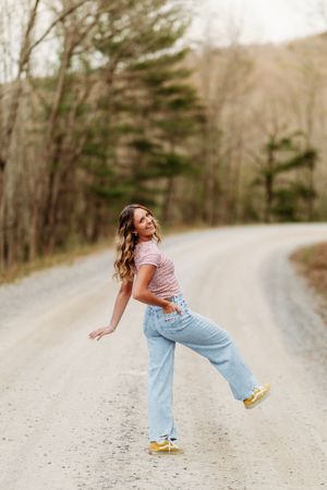 Smiling young woman in pink crop top and wide-leg jeans playfully poses on a scenic gravel road.