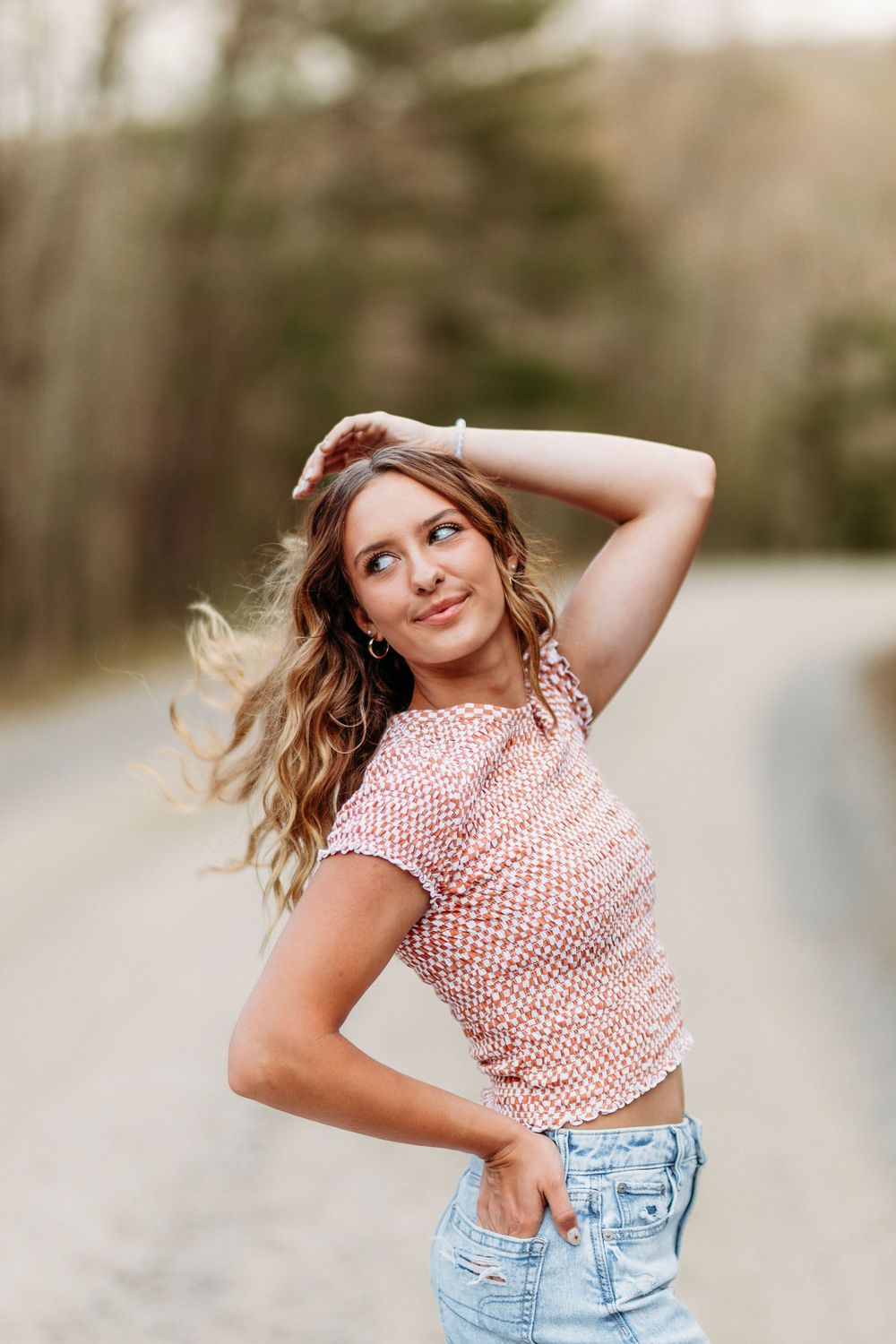 Young woman in pink patterned crop top poses with hand in hair on a blurred outdoor road background.