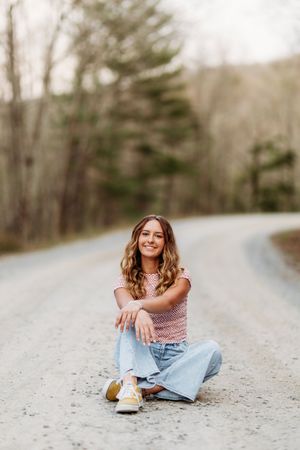 Young woman in pink crop top and wide-leg jeans sits cross-legged on a rural gravel road surrounded by trees.