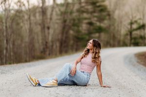 Young woman in pink tank top and jeans sits relaxed on a gravel road surrounded by bare trees.