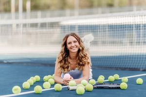 Smiling woman lying on tennis court surrounded by yellow tennis balls.