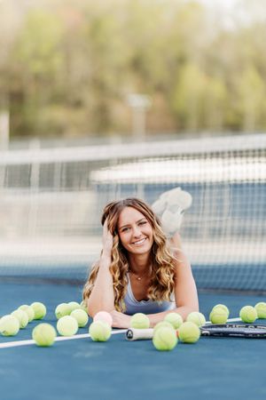 Smiling woman lying on tennis court surrounded by yellow tennis balls.