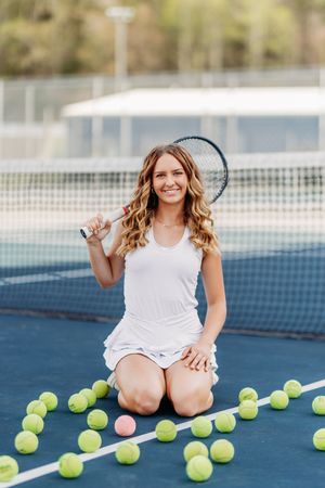 Smiling woman kneeling on tennis court with racket and tennis balls.