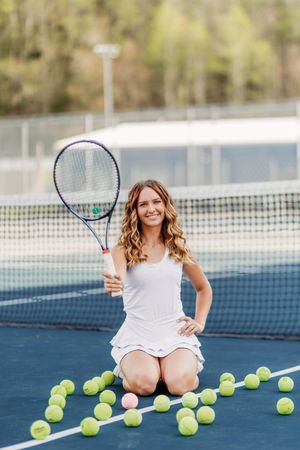 Female tennis player kneeling on court holding racket surrounded by yellow and pink tennis balls.
