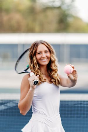 Smiling female tennis player extending a pink ball toward camera while holding racket on court.