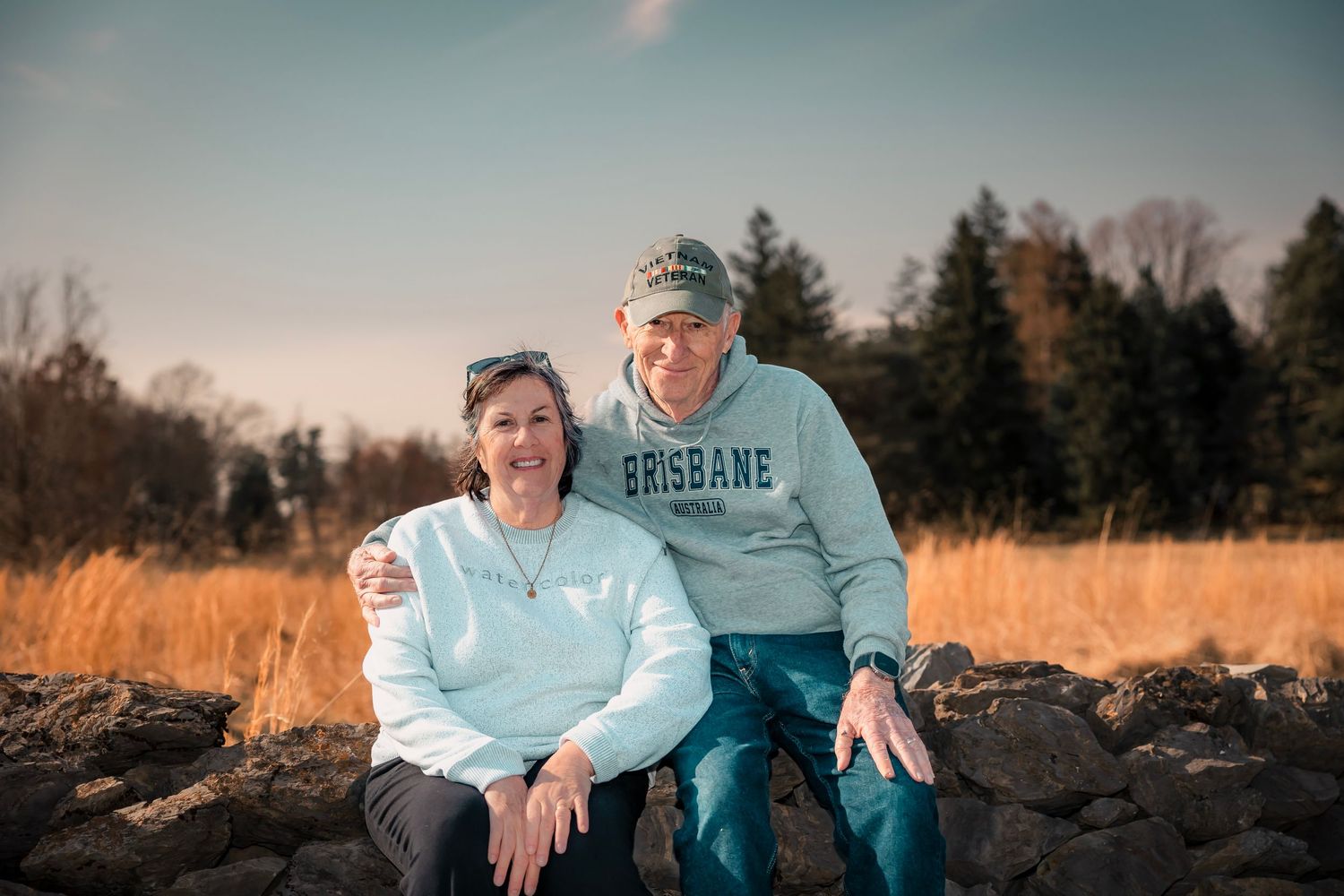 Two people sit together on rocks with golden grass and evergreen trees in the background on a bright autumn day.
