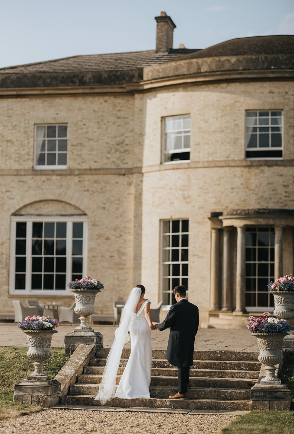 A couple ascends elegant stone steps at a grand Georgian manor house with decorative garden urns and classic architecture.