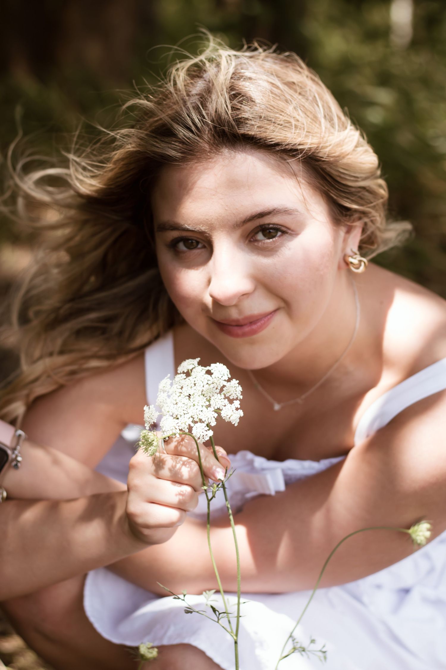 Young woman in white dress holding white wildflowers, smiling outdoors with wind-blown hair in natural setting.
