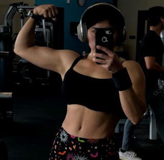 A person takes a selfie in a gym mirror while flexing their arm, wearing a sports bra and patterned workout pants.