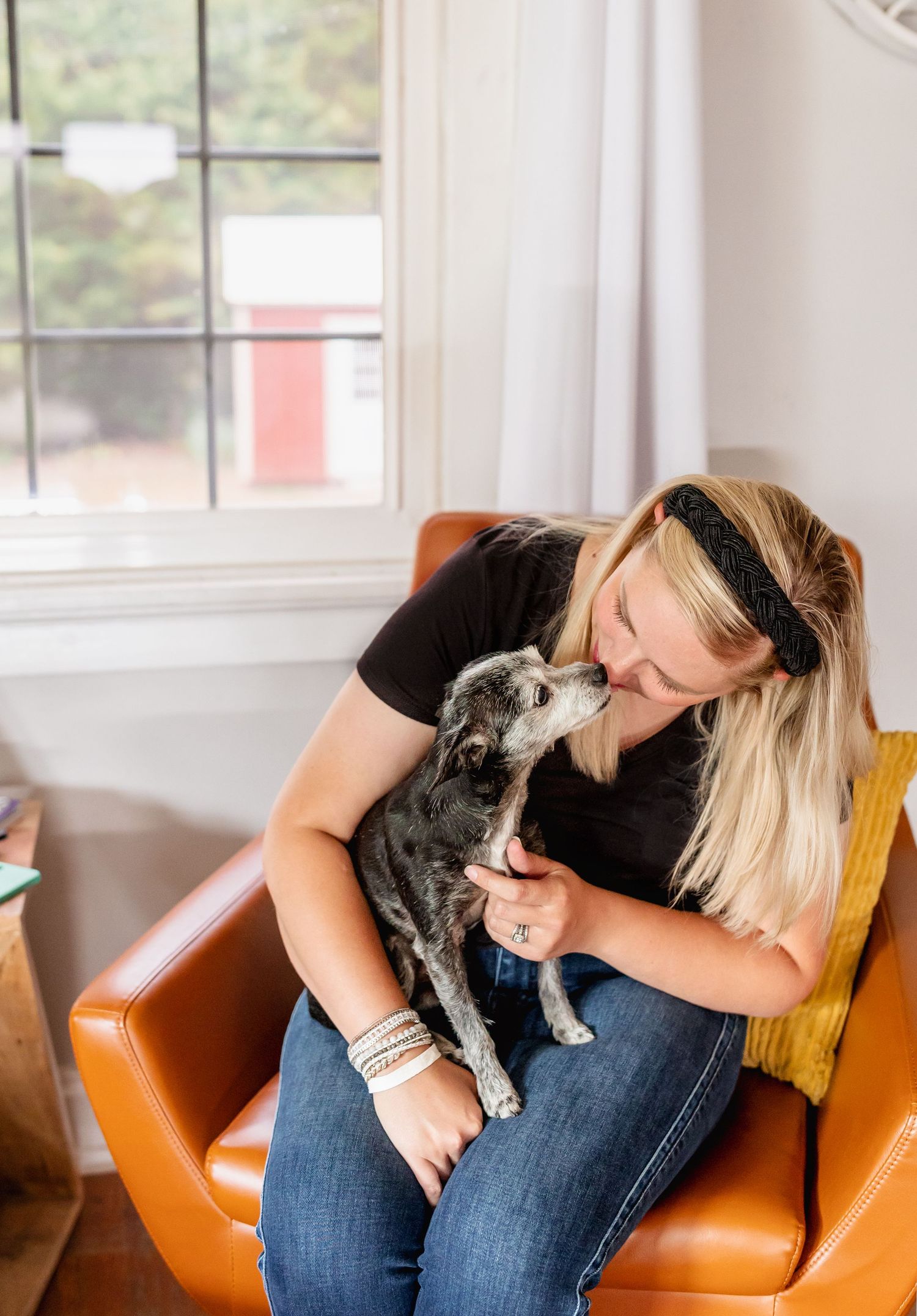 Person relaxing in orange leather chair with small dog in bright, airy room.